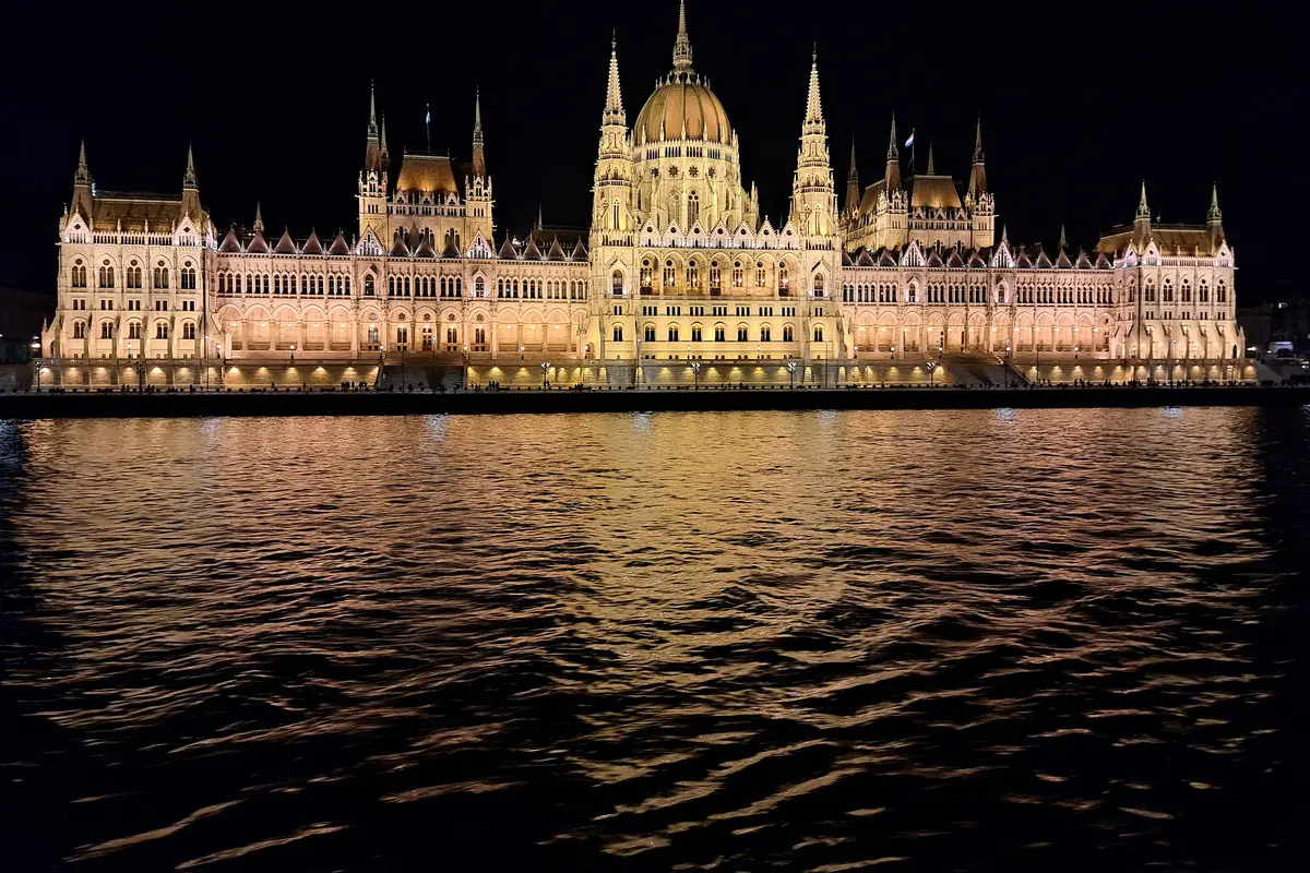 Dlouhé průčelí maďarského Parlamentu nasvícené v noci a odrážející se v temném Dunaji.