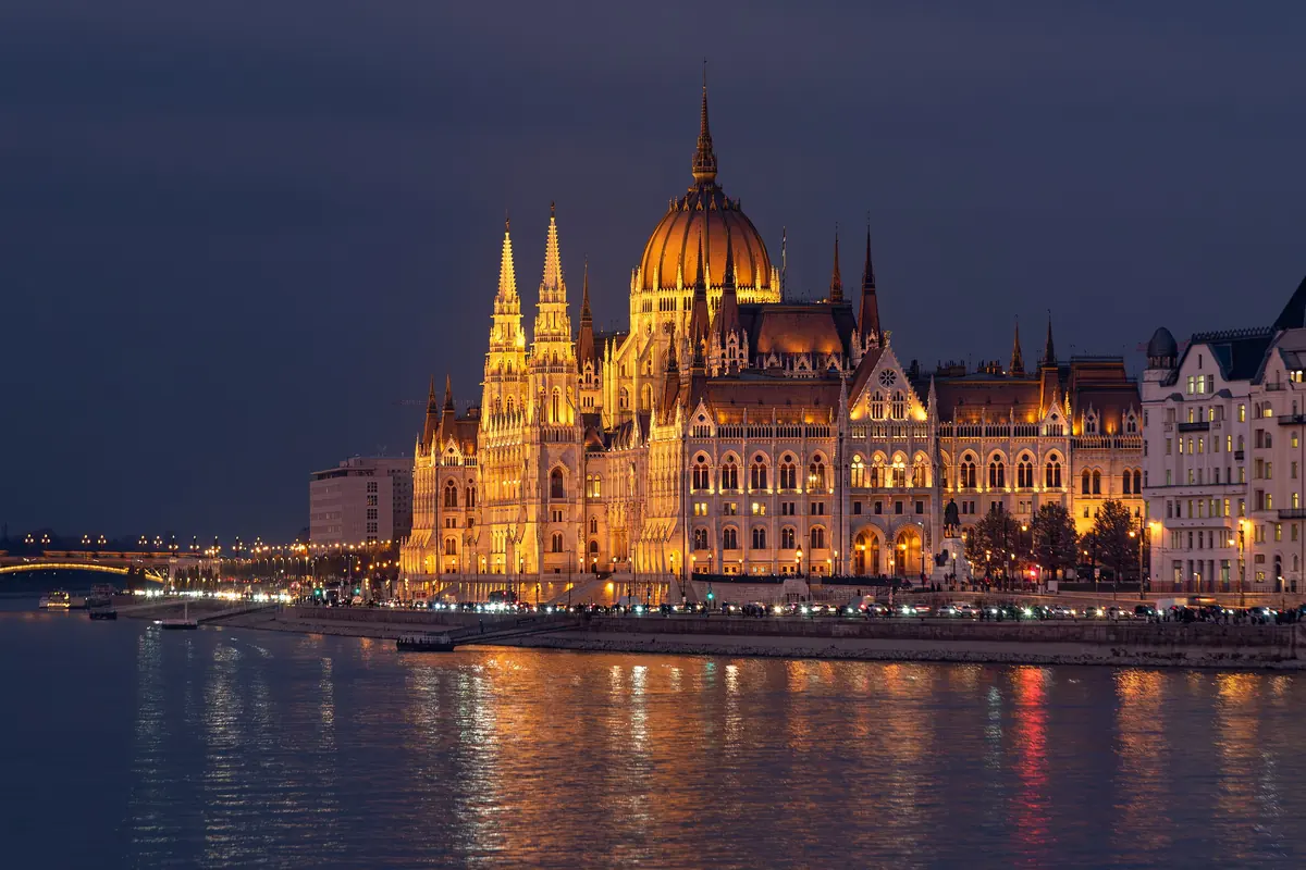Maďarský parlament v soumraku, jeho kopule zlatě nasvícená nad Dunajem, vlevo slabě viditelný Margitin most.