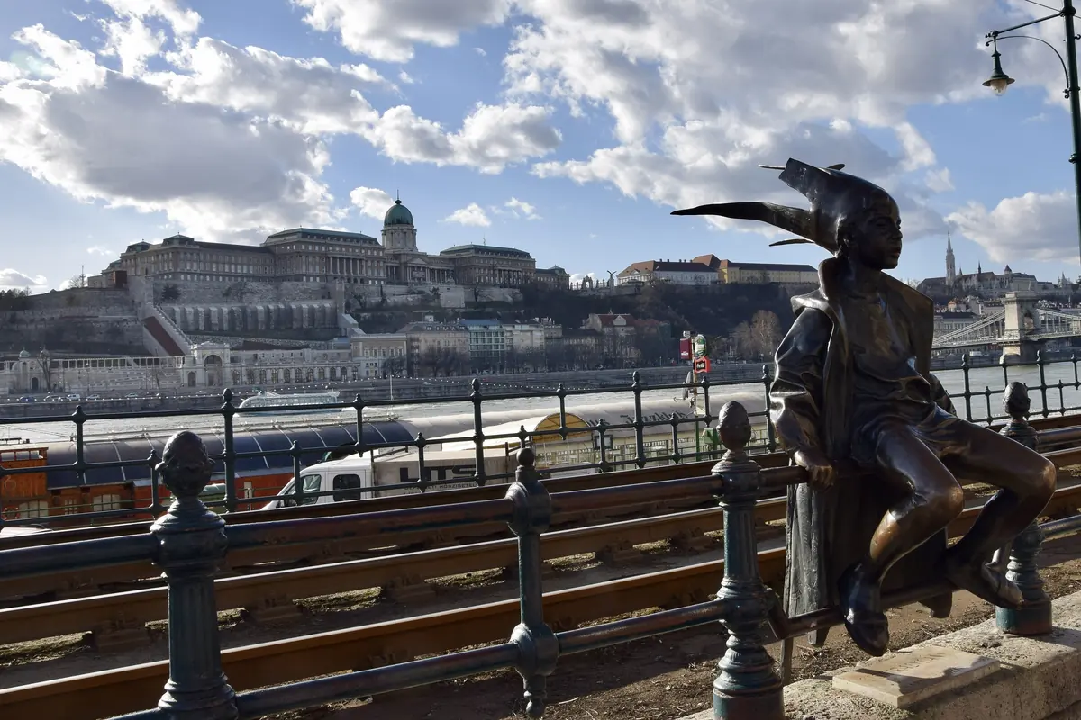 Socha Malé princezny sedící na zábradlí dunajského nábřeží, s palácem Budínského hradu na kopci za řekou.